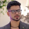 Kashif Aslam wearing glasses, styled in a modern grey suit at the Kashees salon, surrounded by elegant floral decorations.