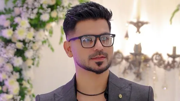 Kashif Aslam wearing glasses, styled in a modern grey suit at the Kashees salon, surrounded by elegant floral decorations.